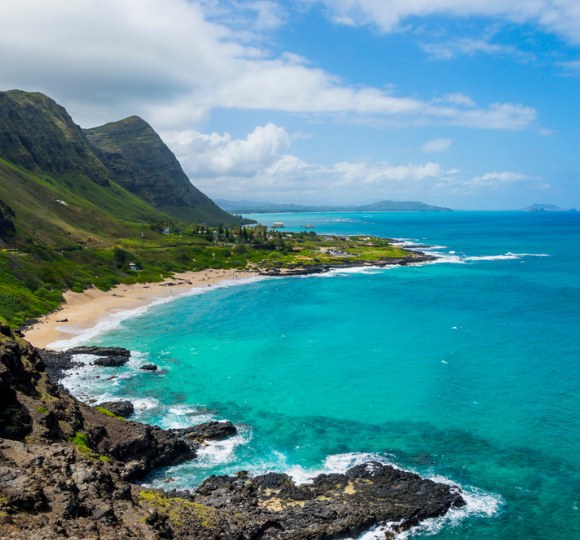 Clear blue ocean water along Honolulu’s shoreline with gentle waves, sandy beach, and lush green mountains in the background
