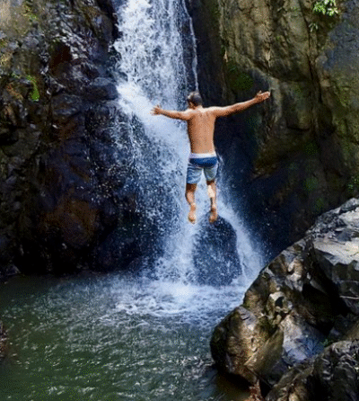 Man jumping into natural pool beneath a waterfall surrounded by lush greenery