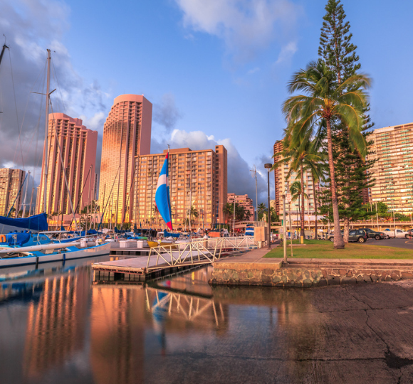Honolulu marina with sailboats docked in calm blue water and city skyline and mountains in the background.